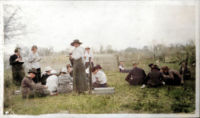 Women and men eating, seated or standing on the grass near a pasture's barbed wire fence, wearing 1918 attire, hats.